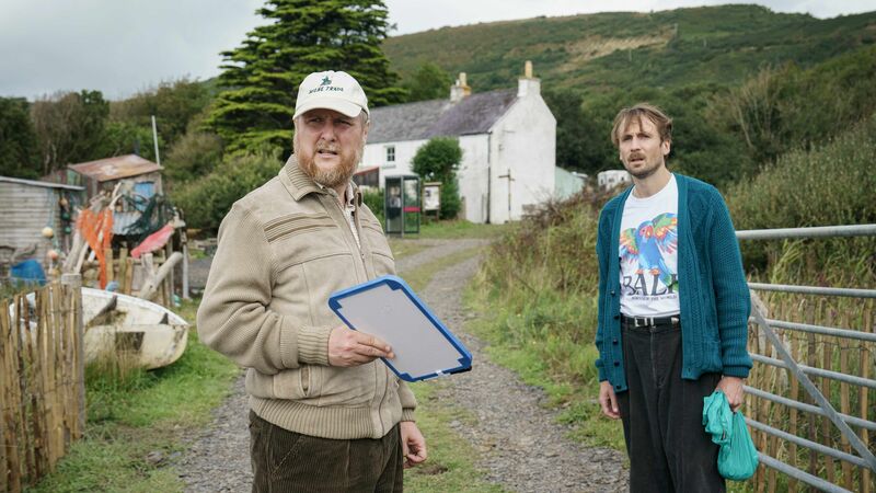 Two men stand outside a country house, one is holding a clipboard, and both look surprised by something they can see off screen.