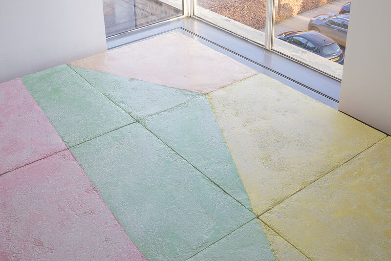 A floor based work is shown in gallery one. The walls are white and the gallery floor is painted grey. On top of this, a sculptural floor is lain in blocks, raising the floor over most of the gallery. This work is brightly coloured, with pastel blue green, yellow orange and pink, in abstract shapes.
