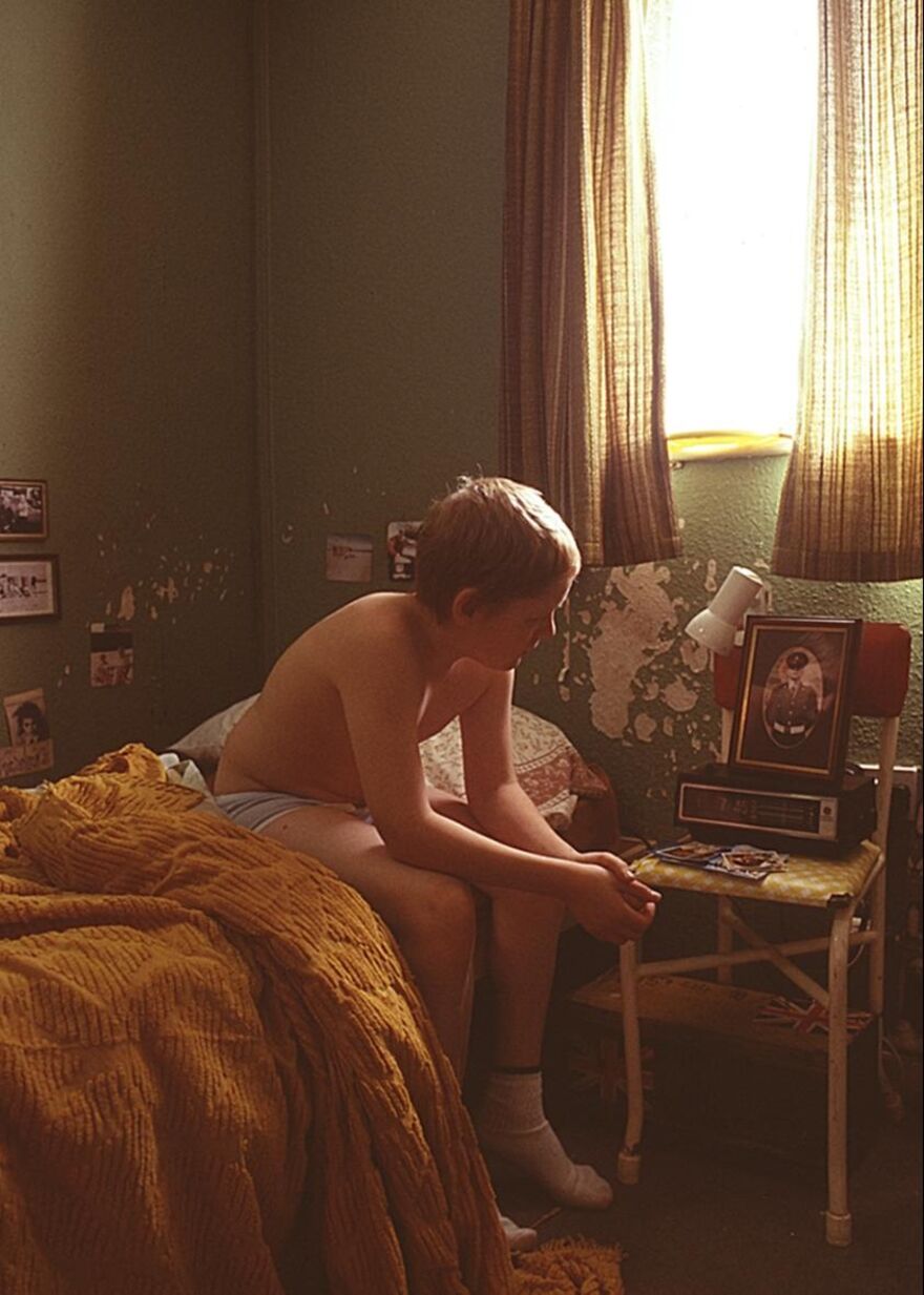A boy sits on the edge of his bed in a 1980s bedroom, looking at an old photograph.
