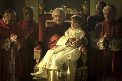 A young boy in a white dress sits on a priest's knee. The priest is sitting on a golden throne. There are other priests surrounding him.
