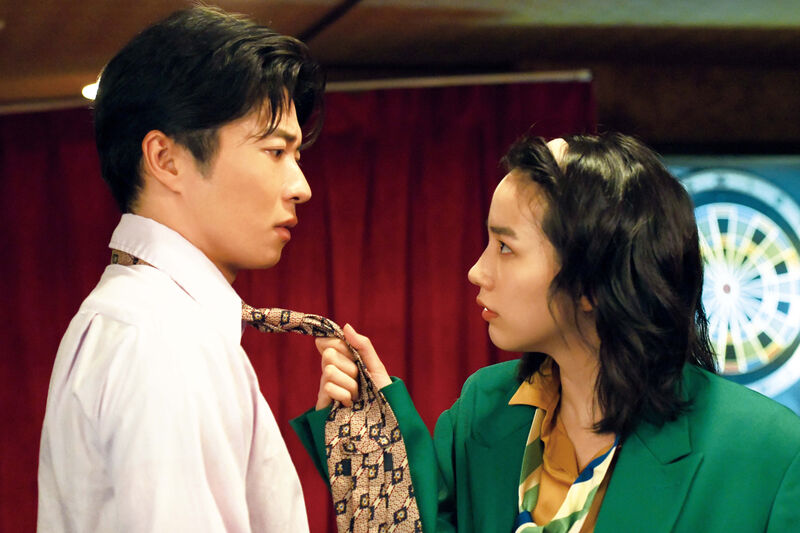 A woman in a baggy green suit holds a man by his tie and looks him in the eye.