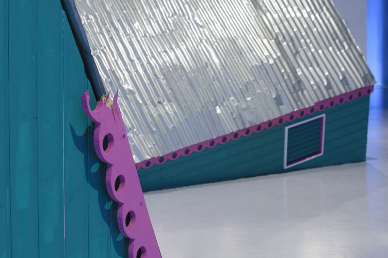 An installation image of gallery two showing a green structure like a building with a corrugated roof and a pink trim, as though it is sinking into the floor.