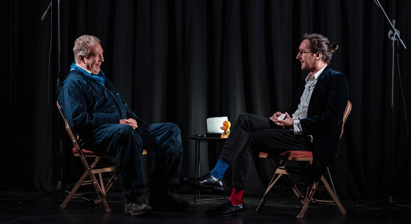 Two men sit opposite each other on stage, talking