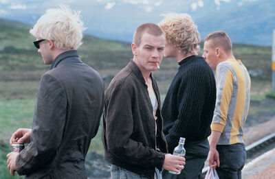Four young men stand outside in the countryside. One has a cigarette in his mouth.