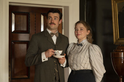 Two people in period clothing stand side by side in an ornate interior, with one holding a floral teacup and saucer beside a wooden door, dark wall panelling, a framed painting, and decorative furniture.