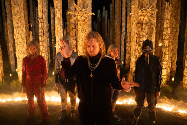 A blonde man in a track suit wearing chains stands in front of a temple made out of bones, dancing.