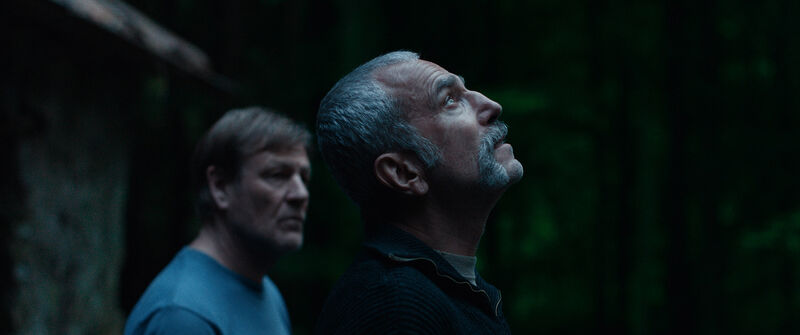 A man in a dark room looks up at the sky, while another man watches him