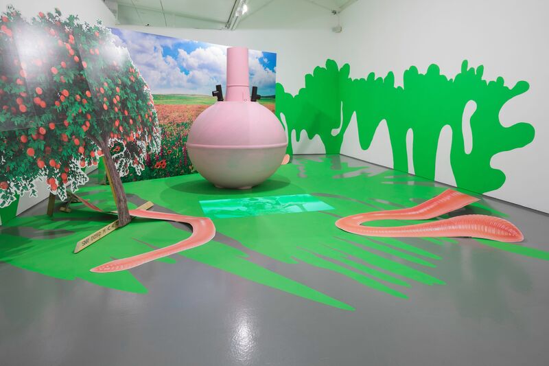 Heather Phillipson's exhibition - a large pink circular structure sits in DCA Galleries. It is sitting on a floor covered in green paint, and there are green paint splotches on the walls. Behind the structure, is a large printed photograph of a blue sky with a green field. There is a cut-out photograph of a green tree with red flowers in front of the pink structure, and two large cut-out photographs of worms.