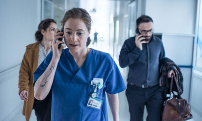 A nurse in a blue uniform walks down a corridor talking on the phone.