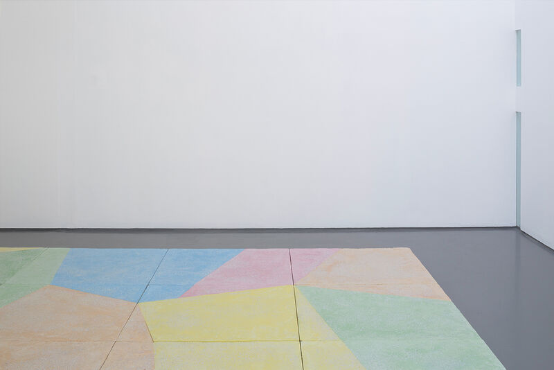 A floor based work is shown in gallery one. The walls are white and the gallery floor is painted grey. On top of this, a sculptural floor is lain in blocks, raising the floor over most of the gallery. This work is brightly coloured, with pastel blue green, yellow orange and pink, in abstract shapes.