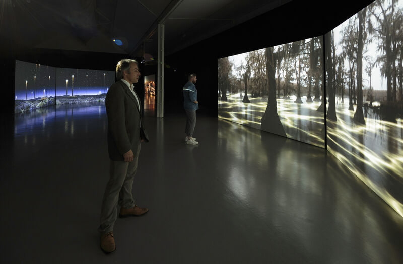 Image shows visitors watching a film in the gallery projected over two large screens to the right of the photograph. The film shows a landscape with a green glowing swamp and trees. Behind the visitors, to the left, can be seen another film projected over two screens, with a blue glowing landscape with bright lights in a dark sky.