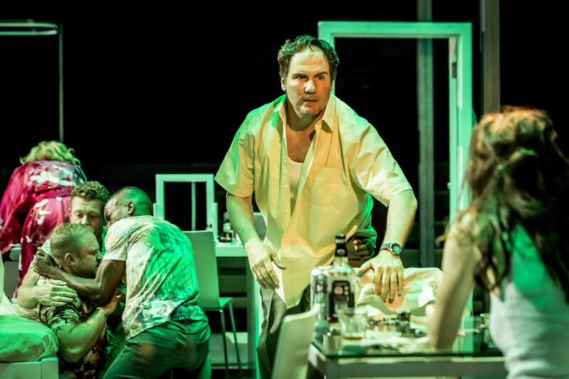 A man in a white shirt leans aggressively over a table, while three men hug in the background.