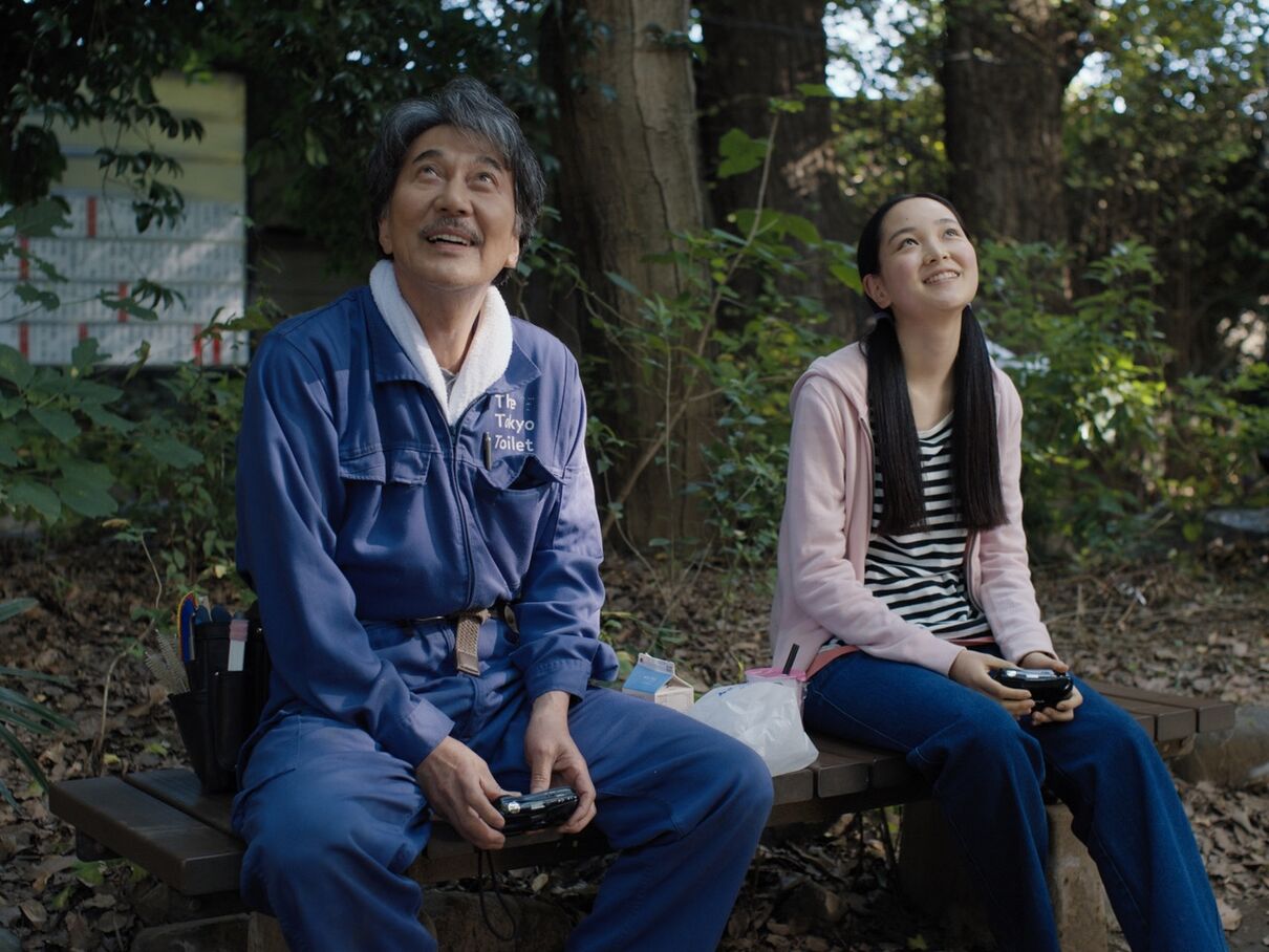 An older man with grey hair, wearing a toilet-cleaning uniform, and a young girl with hair in bunches, sit on a park bench together.