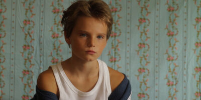 A child with short blonde hair and a white vest sits in front of flowery wallpaper.