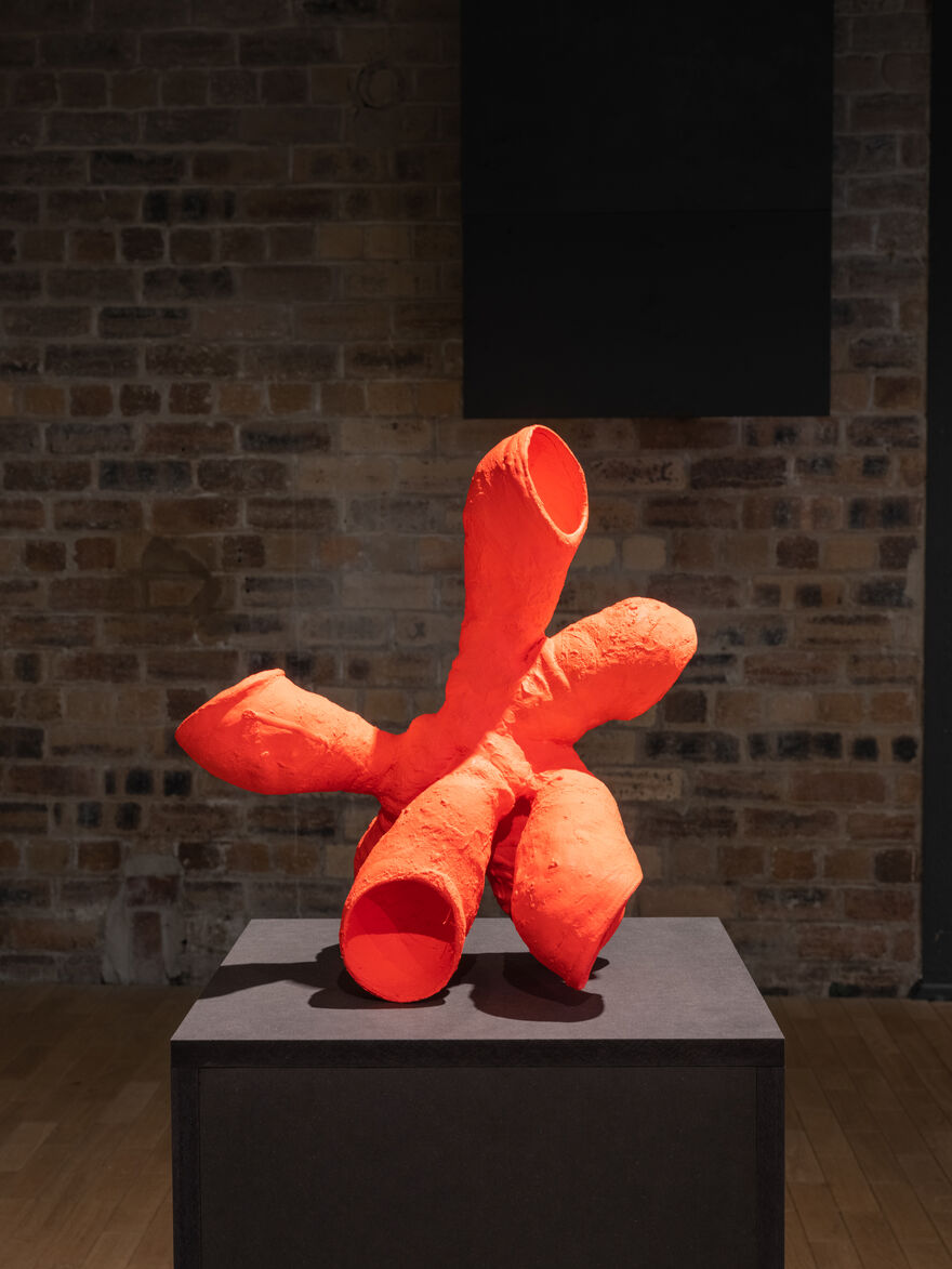 An orange sculptural object sits on a pinlth in a gallery space with a brick wall