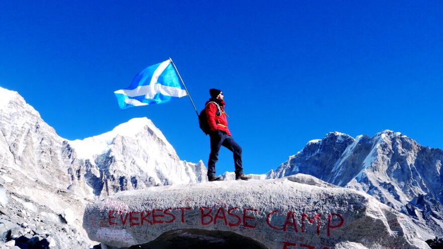 A man stands on top of a boulder in the Himalaya with Everest Base Camp painted on it. He carries a Scottish Flag