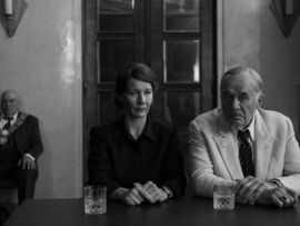 A woman and an older man sit facing the camera. A man in uniform sits in the corner behind them