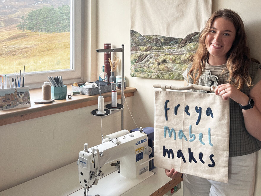 Freya Turner in her studio holding a fabric banner which says 'freya mabel makes'