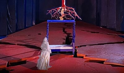 A woman on a stage in a white robe, a cracked red floor beneath her, looks at a blue bedpost and a red tree
