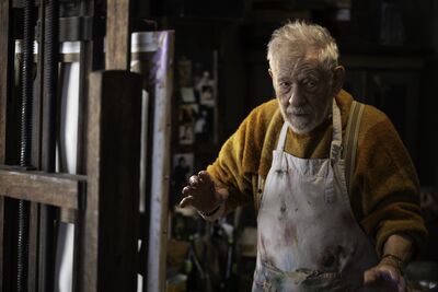 Ian Mckellan is stood in front of an easel wearing a paint stained apron and yellow jumper