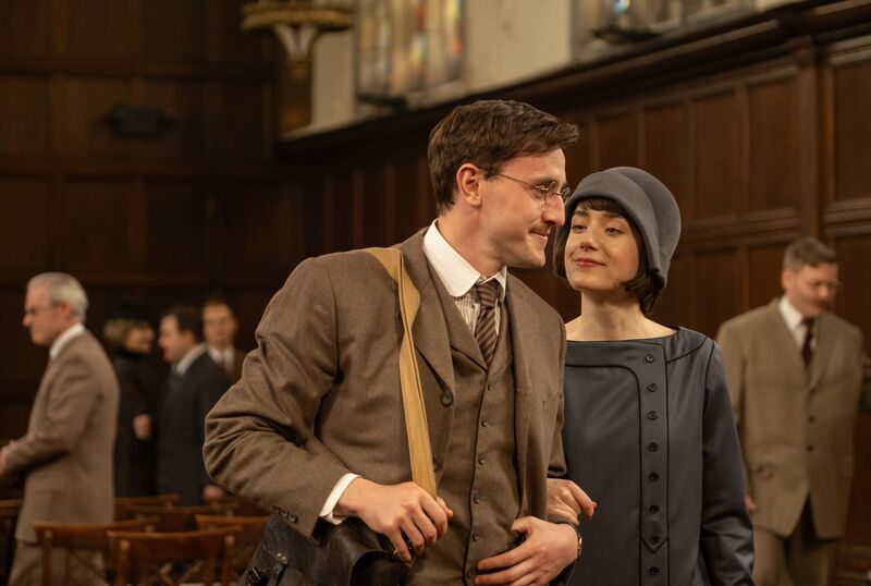 A man wearing a three-piece brown suit and glasses smiles next to a woman wearing 1920s clothes.