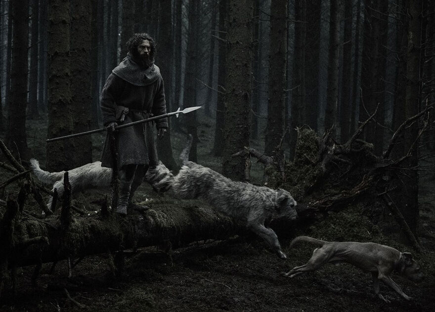 A man stands carrying a lance as three dogs run past him