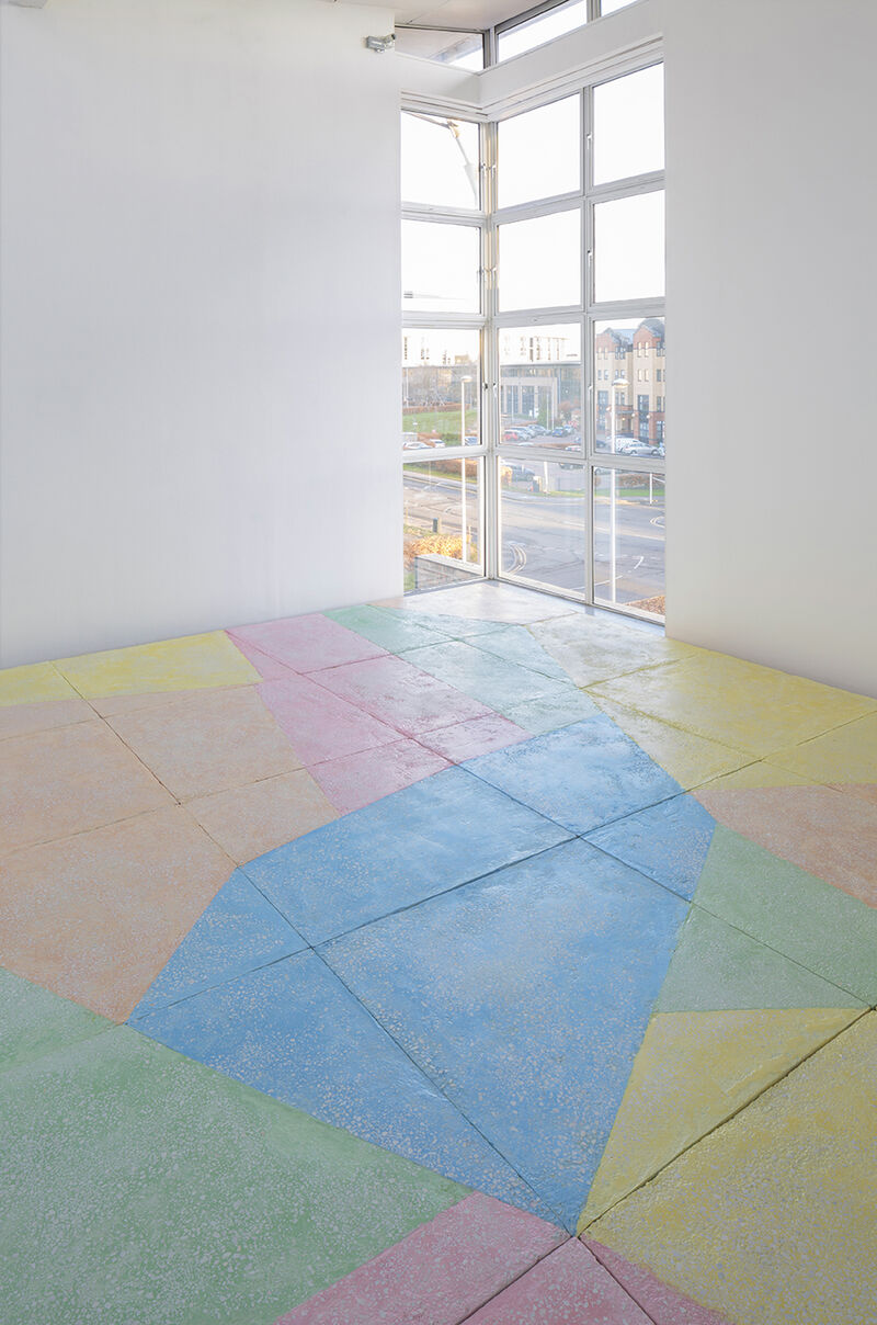 A floor based work is shown in gallery one. The walls are white and the gallery floor is painted grey. On top of this, a sculptural floor is lain in blocks, raising the floor over most of the gallery. This work is brightly coloured, with pastel blue green, yellow orange and pink, in abstract shapes.