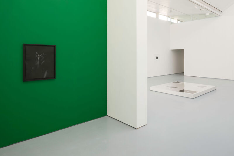 Installation view of Gallery 1 showing a green wall with a dark photograph to the right hand side, and in the left mid ground a low display case containing photographs.