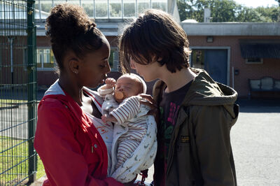 A young black girl holds a small baby with a young while male with curtain hair. They both look at the baby lovingly