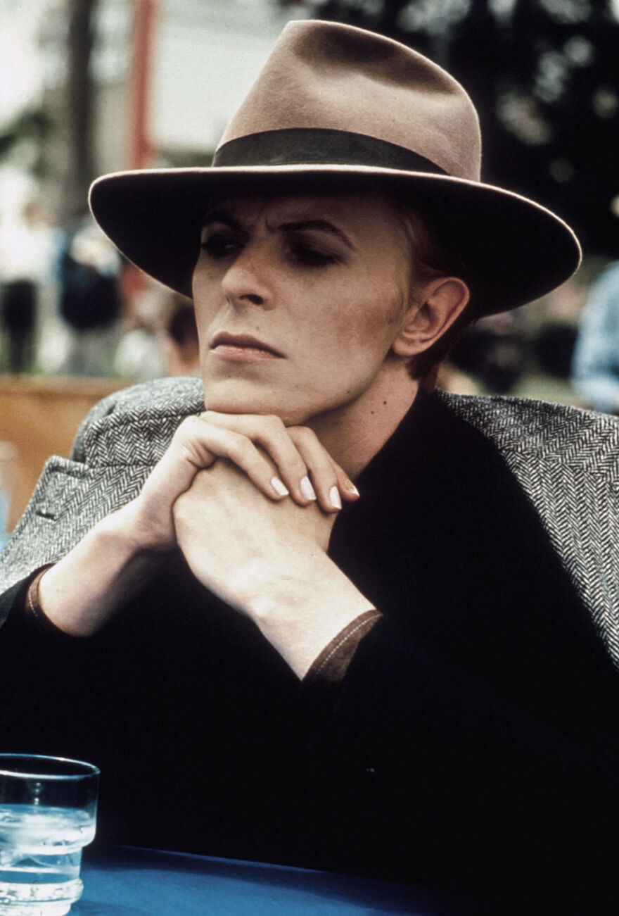 A man sits at an outdoor table wearing a fedora