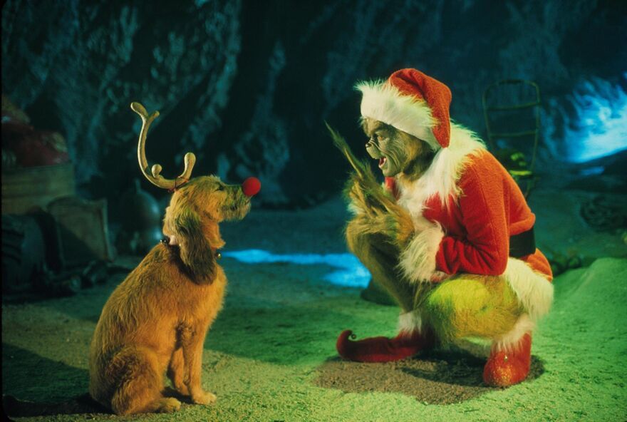 A green creature in a santa outfit crouches next to a dog wearing antlers and a red nose