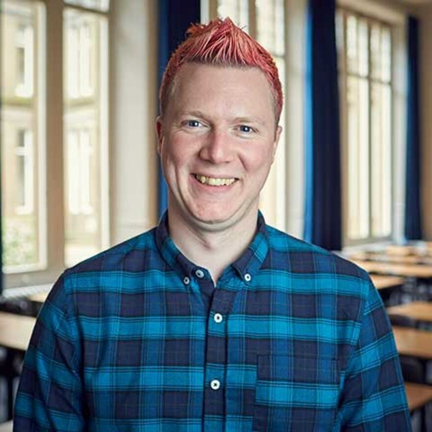 A headshot of Paul Robertson, a man with pink hair wearing a blue shirt
