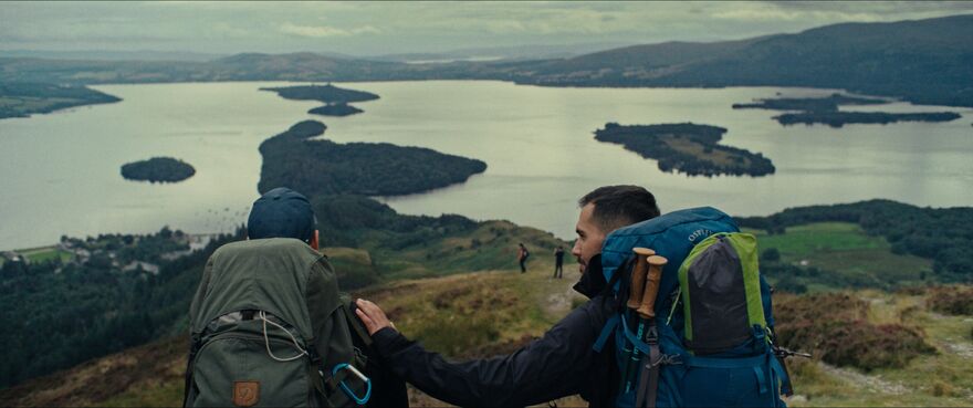 Two men in hiking gear sit on a hill looking out at a loch
