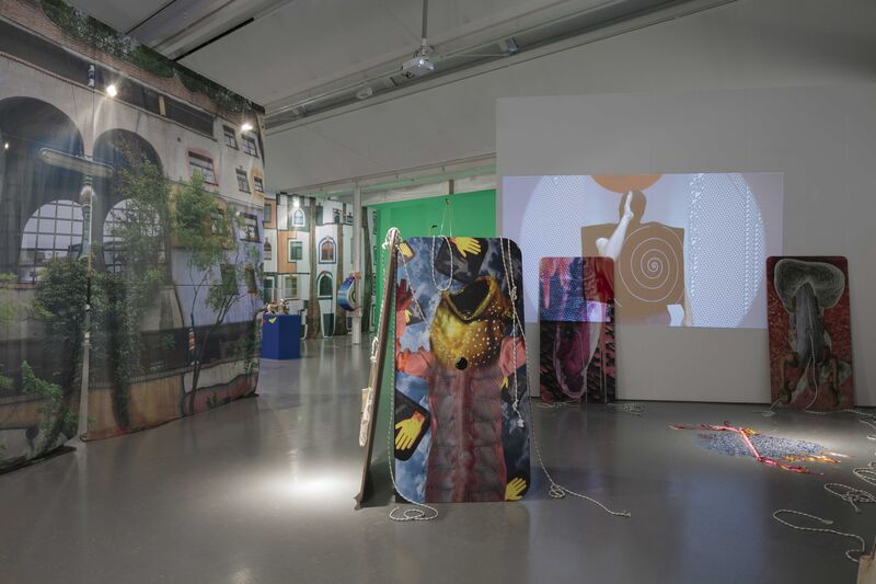 An image from 'Shonky: The Aesthetics of Awkwardness' shows multiple colourful canvases with pictures of fish on them. Next to this, is a large image of a colourful building printed on fabric. In the background, a projector shows a figure wearing a black box.