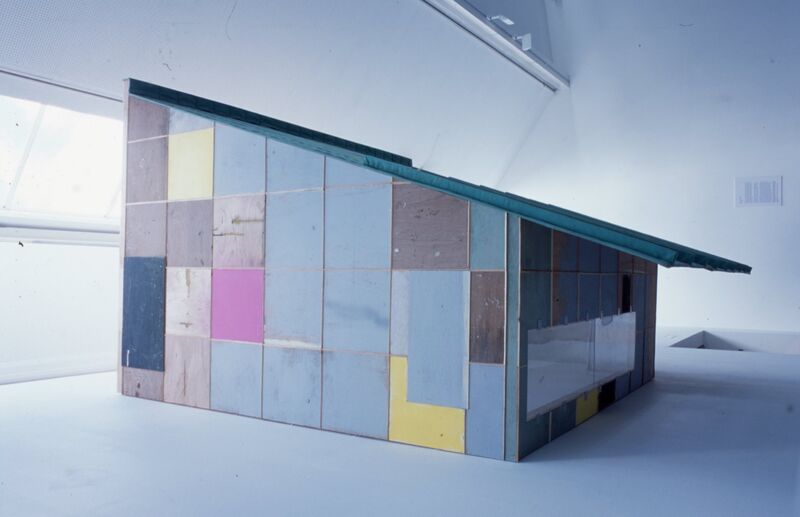 A diagonal, shed-like building made up of coloured panels. This sits in a white gallery space.