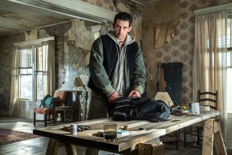 A man is stood in a house that is falling apart, he is packing a bag at a dining table.