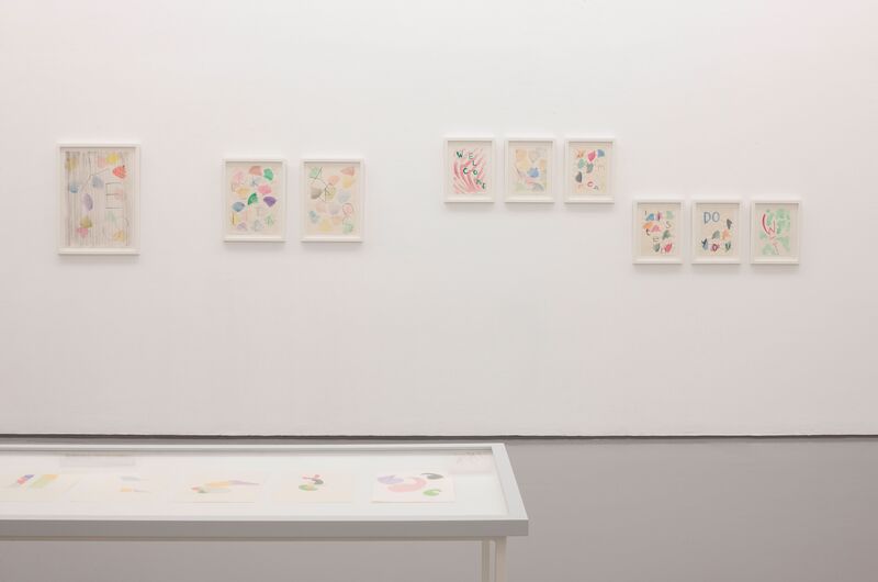 An image from Katy Dove's exhibition shows abstract, colourful prints on a white wall. In the foreground, there is a white table which also displays colourful drawings.