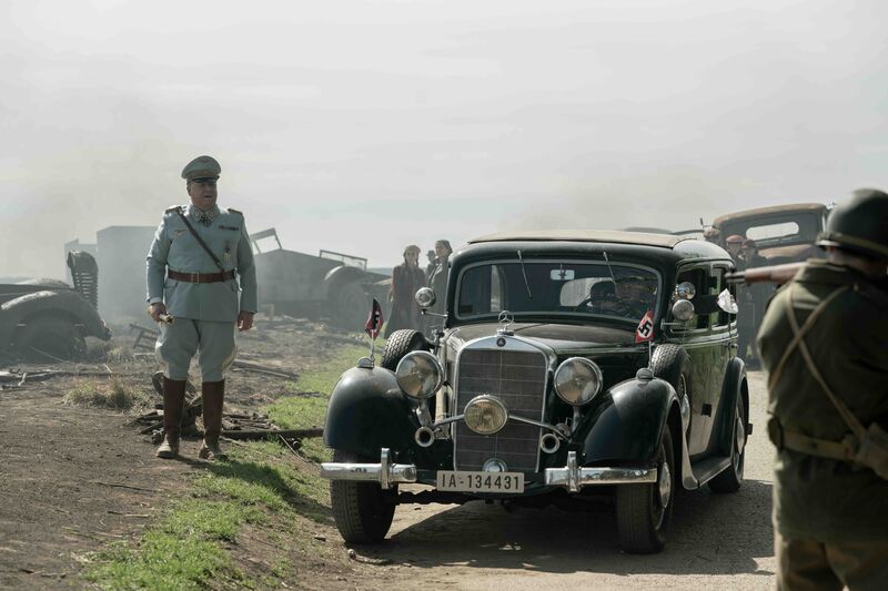 A Nazi official stands next to a car decorated with Nazi insignia. 