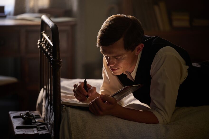 A man in a waistcoat lies on his bed, writing a letter.