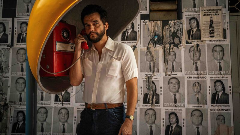 A man talks in a phonebooth while standing against a wall with mugshots on it