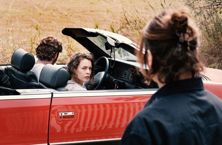 A woman in a red car looks at an older woman dressed in black nearby.