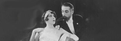 A woman dressed in a flapper outfit smokes a pipe, next to a man in a suit and white bowtie.