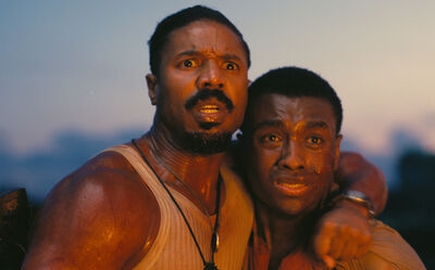 Two men stand, one with their arm around the other, looking terrified while they stare at something.