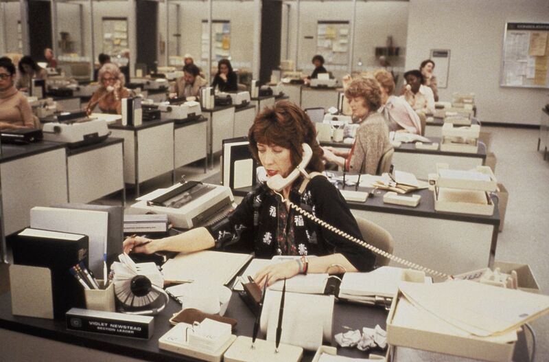 A woman in a busy office talks on a telephone while sitting at her desk