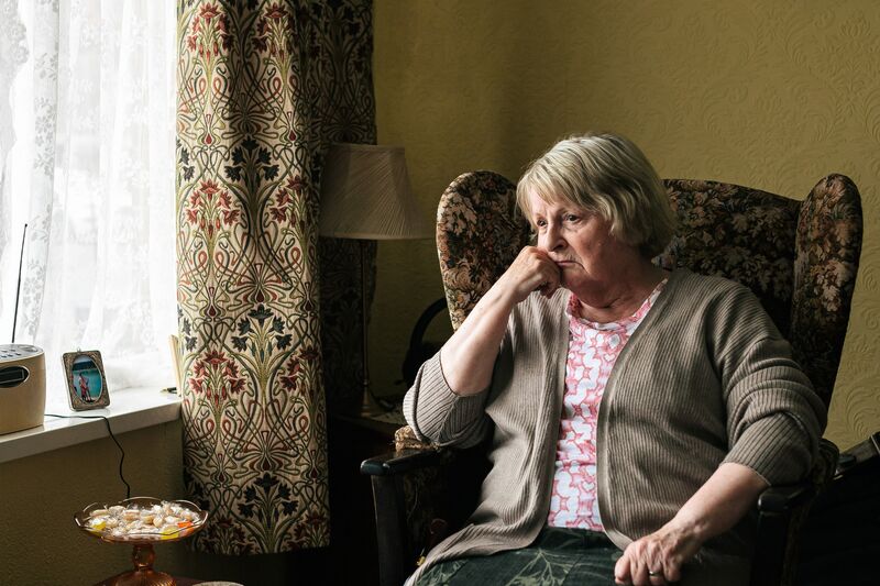 An older woman wearing a cardigan sits on an armchair and looks out the window.