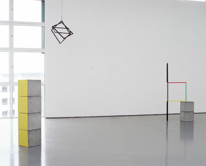 From Camilla Løw's exhibition in DCA. Four grey cubes are stacked on top of each other. One side of each cube is yellow. Black metal frame hang from the ceiling. A sculpture made up of coloured metal frames and grey cubes can be seen in the distance.