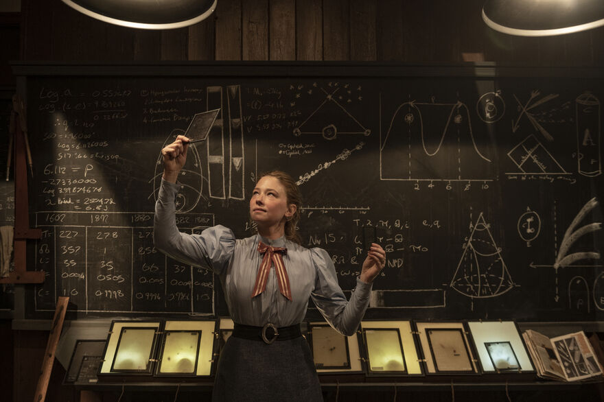 A person in period clothing stands in front of a large chalkboard covered with scientific diagrams, equations, graphs, and notes, holding photographic plates under warm overhead lighting.