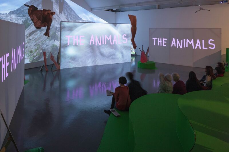 From Heather Phillipson's exhibition. Visitors to DCA Galleries sit on a hill made out of fake grass in DCA gallery. They watch three projector screens, which say 'The Animals' on them in purple writing.