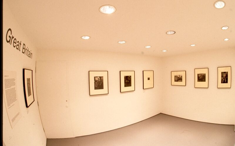 A fish eye lens photo of Small black-and-white photos on white backing hang on a. white wall. Text above the frames says  'Great Britain'
