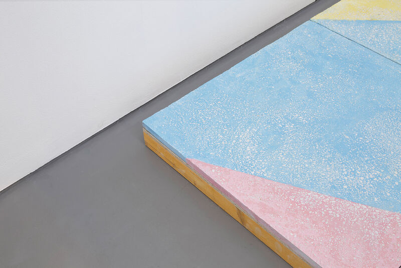 A floor based work is shown in gallery one. The walls are white and the gallery floor is painted grey. On top of this, a sculptural floor is lain in blocks, raising the floor over most of the gallery. This work is brightly coloured, with pastel blue green, yellow orange and pink, in abstract shapes.